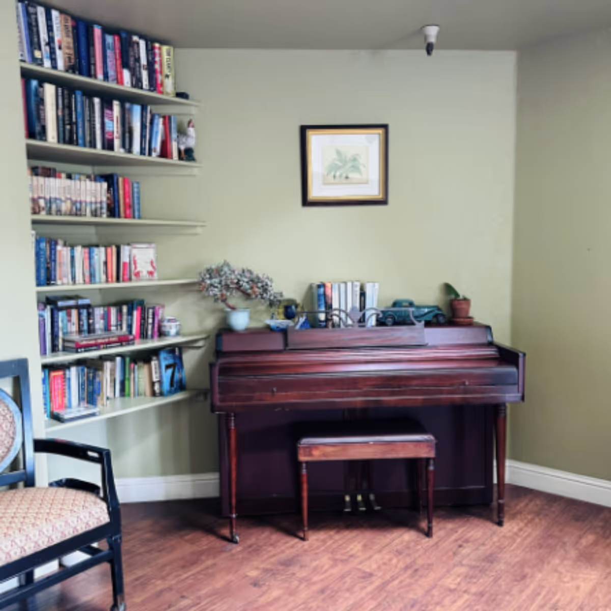 A cozy corner of a room with a wooden piano and matching bench against a light green wall. Above the piano hangs a framed picture of a plant. To the left, there are several shelves filled with books and decorative items. A cushioned chair with a patterned seat is partially visible on the left side. The floor is wooden.