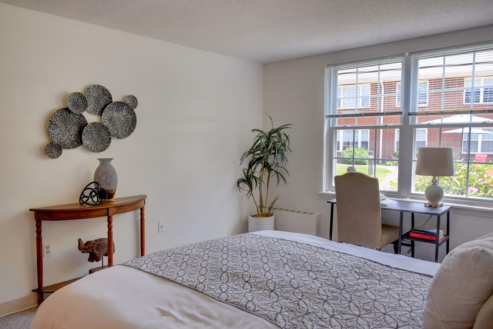 Bright bedroom with a made bed, decorative wall art, small console table, potted plant and a desk by a large window overlooking the courtyard.
