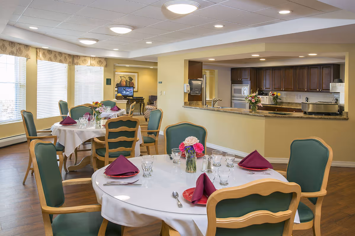 A bright dining room with round tables covered in white tablecloths, each set with glassware, silverware, and folded burgundy napkins. The chairs have wooden frames with green upholstery. In the background, there is a kitchen area with wooden cabinets, a microwave, and a countertop with flowers. Large windows with blinds allow natural light to fill the room.