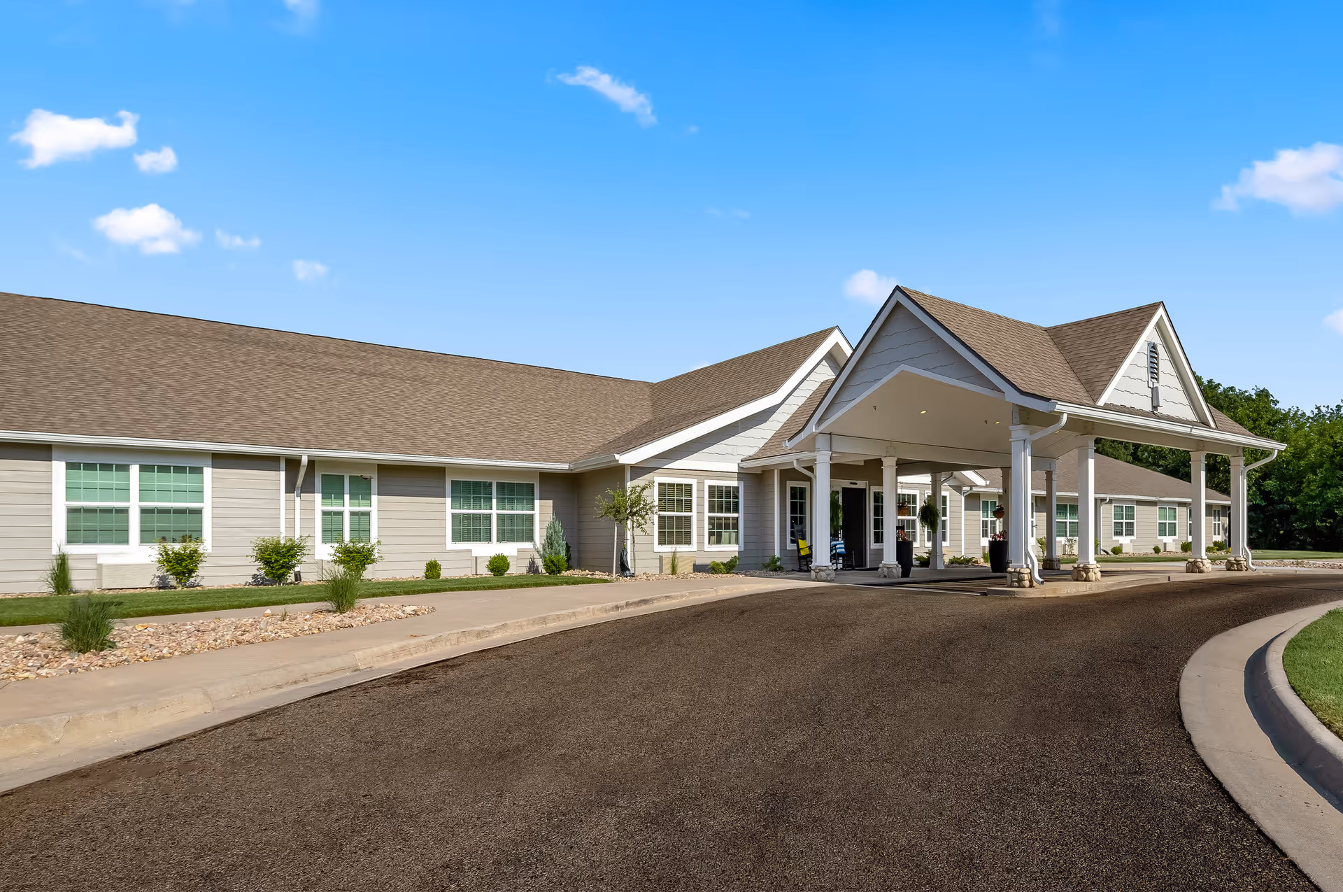 Exterior view of a single-story senior living facility building with a covered entrance driveway, beige siding, multiple windows, and a well-maintained landscape under a clear blue sky.