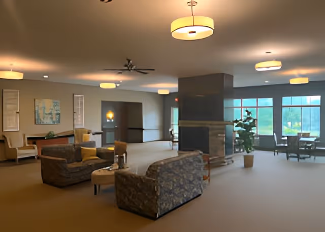 Spacious senior living common lounge with sofas, a central fireplace, tables by large windows, and overhead lights.
