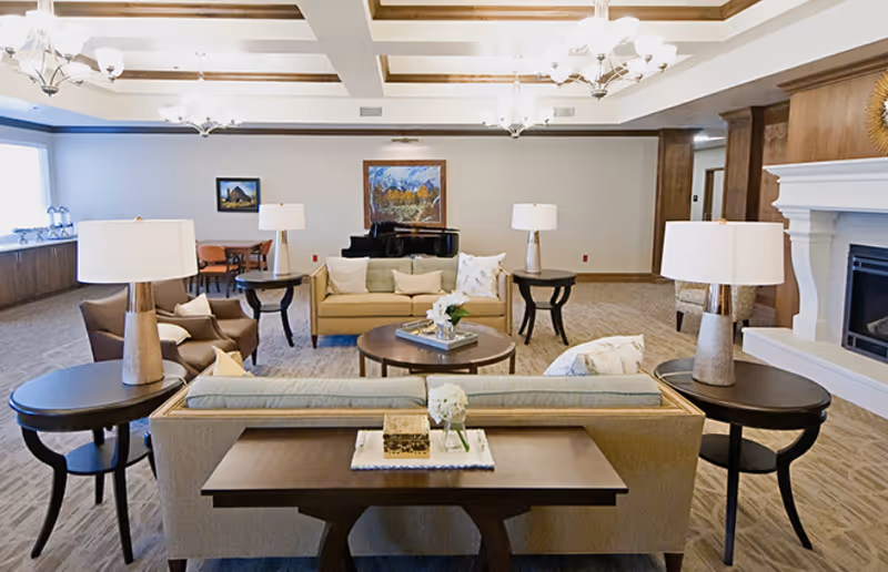 Spacious, well-lit sitting room with sofas, armchairs, side tables and lamps arranged around a central coffee table, a fireplace on the right and a piano in the back.