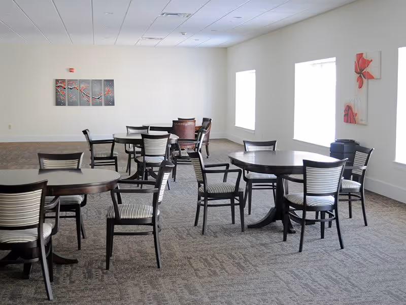 A bright room with multiple round tables and chairs arranged for seating. The room has large windows letting in natural light, light-colored walls, and carpeted flooring. There are two pieces of floral artwork on the walls.