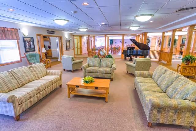 A spacious senior living facility common area with multiple patterned sofas and armchairs arranged around a wooden coffee table with a small plant. The room features beige carpeting, wood trim, framed artwork on the walls, and a grand piano in the background near a dining area with tables and chairs. The ceiling has recessed lighting and ceiling tiles.