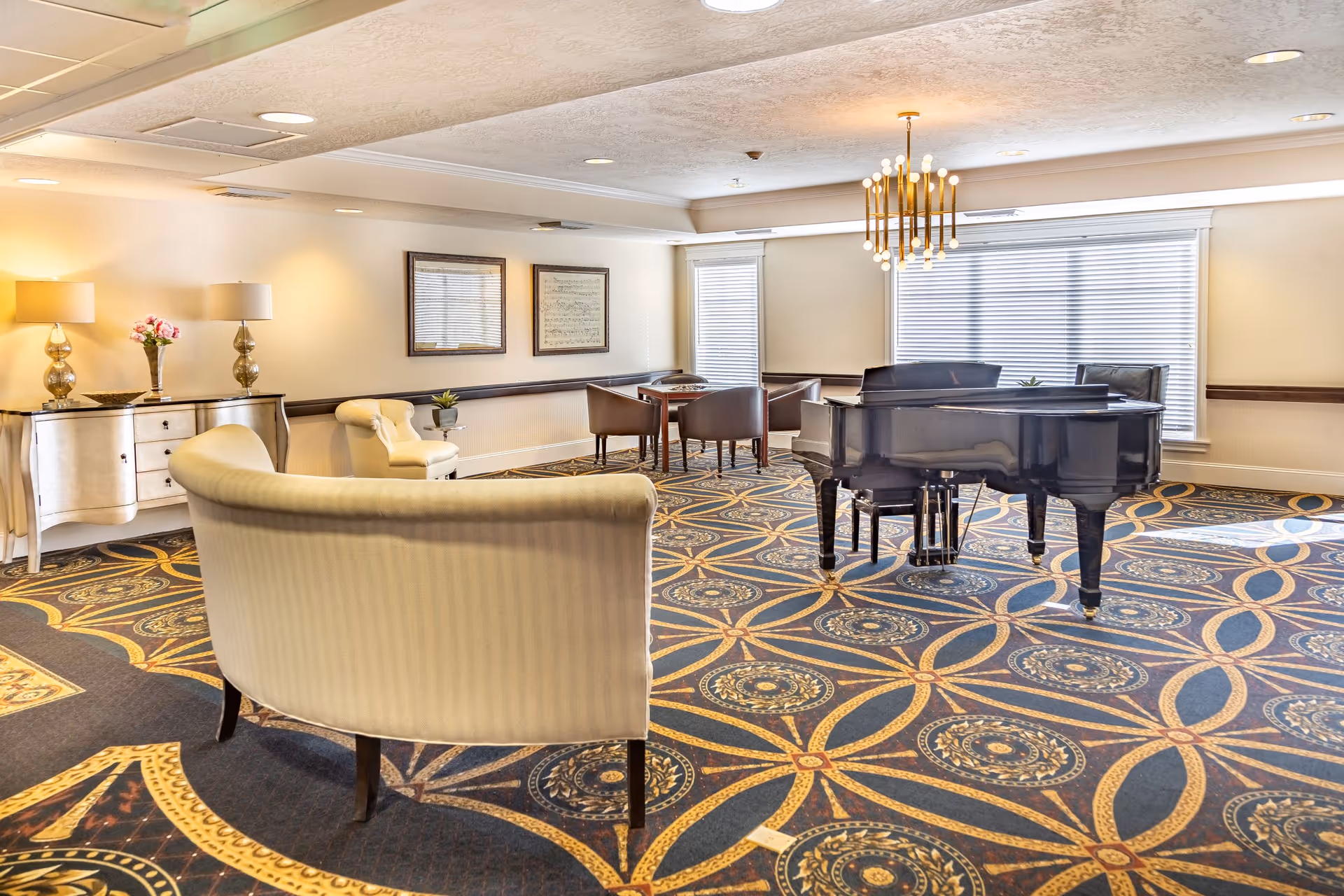 A spacious, well-lit common area with a grand piano in the center, surrounded by comfortable seating including a curved sofa and armchairs. The room features a patterned carpet, a chandelier, framed artwork on the walls, and large windows with blinds allowing natural light to fill the space.