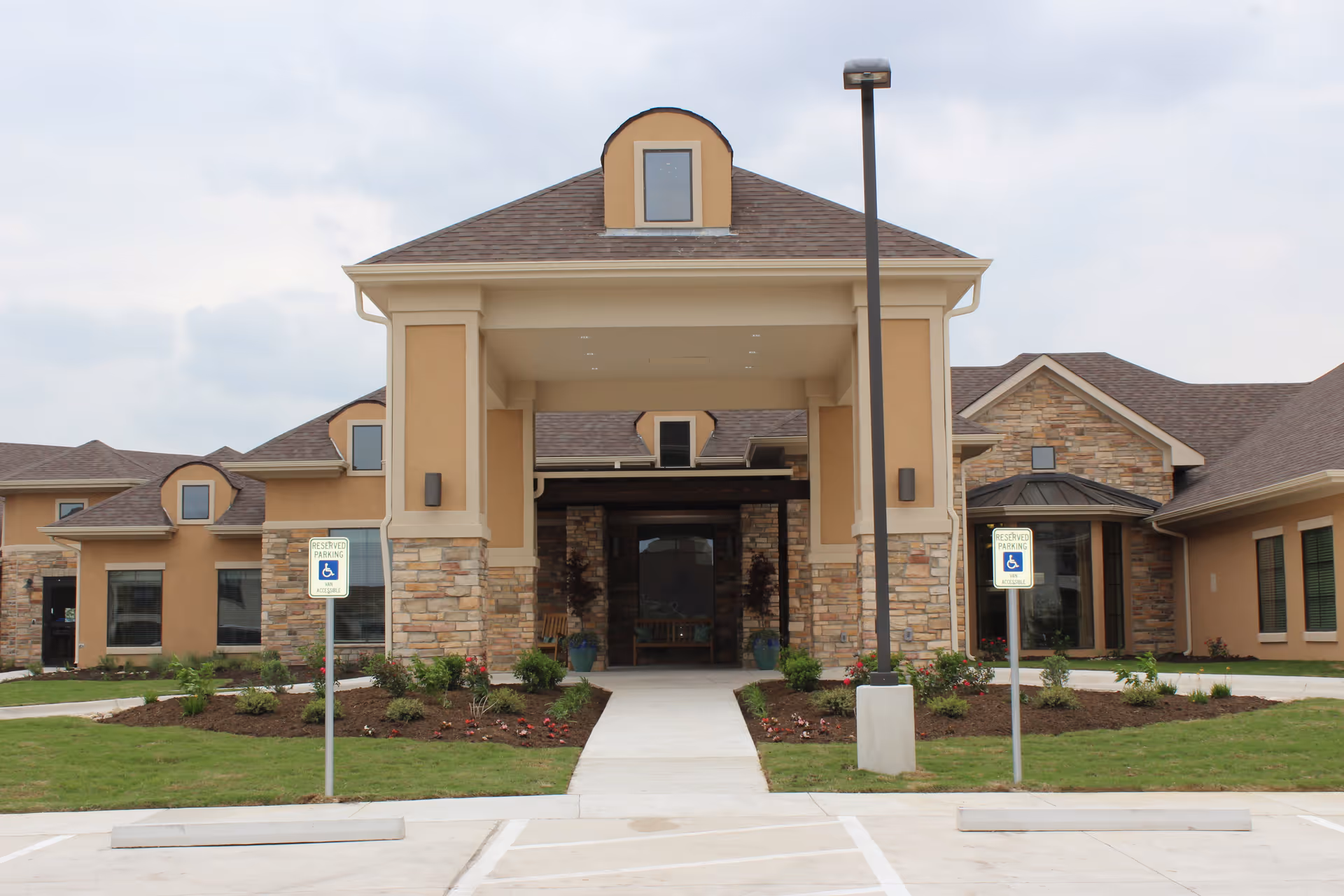 Front exterior view of a single-story building with a covered entrance supported by columns. The building has a combination of stone and beige stucco walls with multiple windows and a brown shingled roof. There are two reserved parking signs for handicapped parking near the entrance, and a landscaped area with grass and small plants in front.
