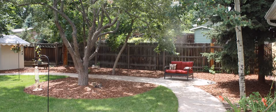 Backyard with curved concrete path, trees, mulch, a wooden fence, and a red cushioned outdoor chair.