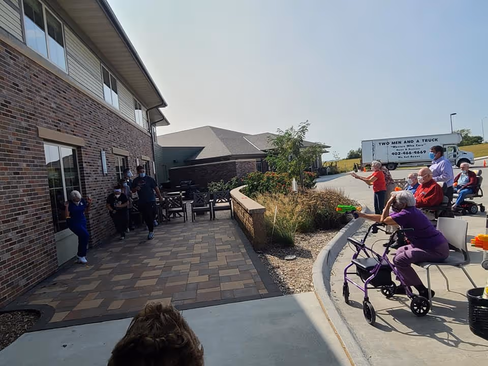 Residents and staff gathered outside a brick assisted living building on a patio and driveway, some seated and some aiming water guns.