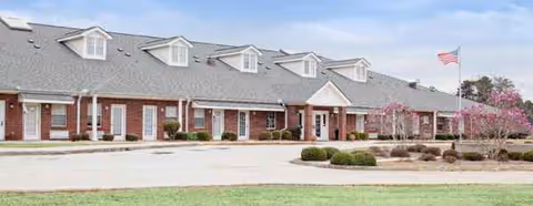 Brick senior living building with dormer windows, flag, and well kept landscaping.