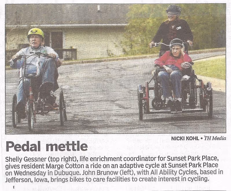 Two people riding adaptive cycles on a paved path. On the left, a man wearing a yellow helmet pedals a three-wheeled cycle. On the right, a woman in a wheelchair-style adaptive cycle is being pushed by another woman wearing a black helmet. The background shows a building and greenery.