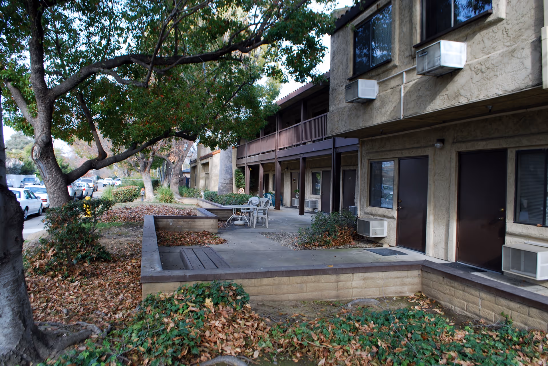 Outdoor view of Leisure Manor Apartments showing a two-story building with beige stucco walls, brown doors, and window air conditioning units. There is a concrete patio area with chairs and a table, surrounded by low brick walls and landscaped with trees and bushes with fallen leaves.