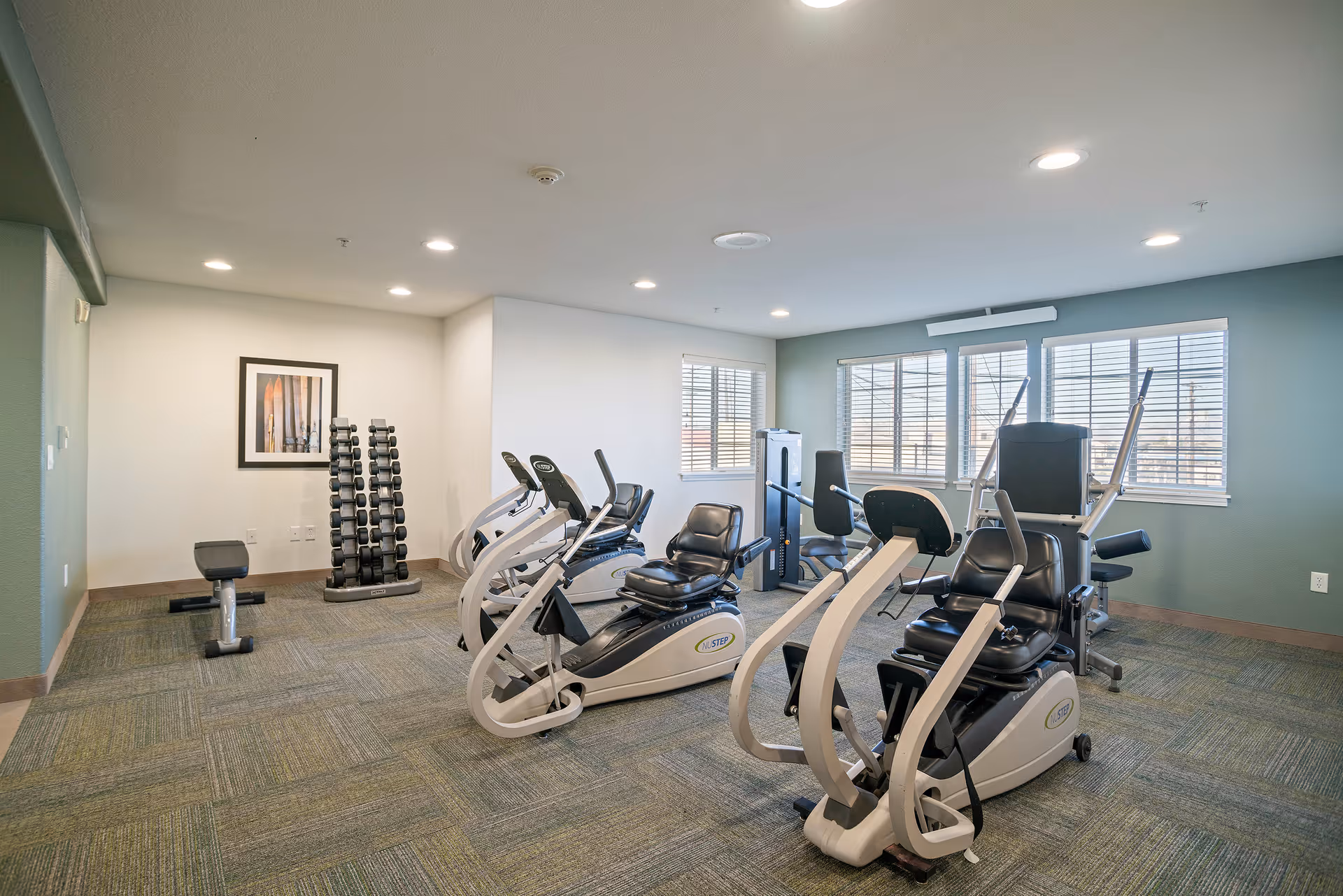 A well-lit exercise room with several pieces of fitness equipment including recumbent bikes and weight machines. There is a rack of dumbbells against the wall and a framed picture hanging above it. The room has large windows with white blinds letting in natural light.