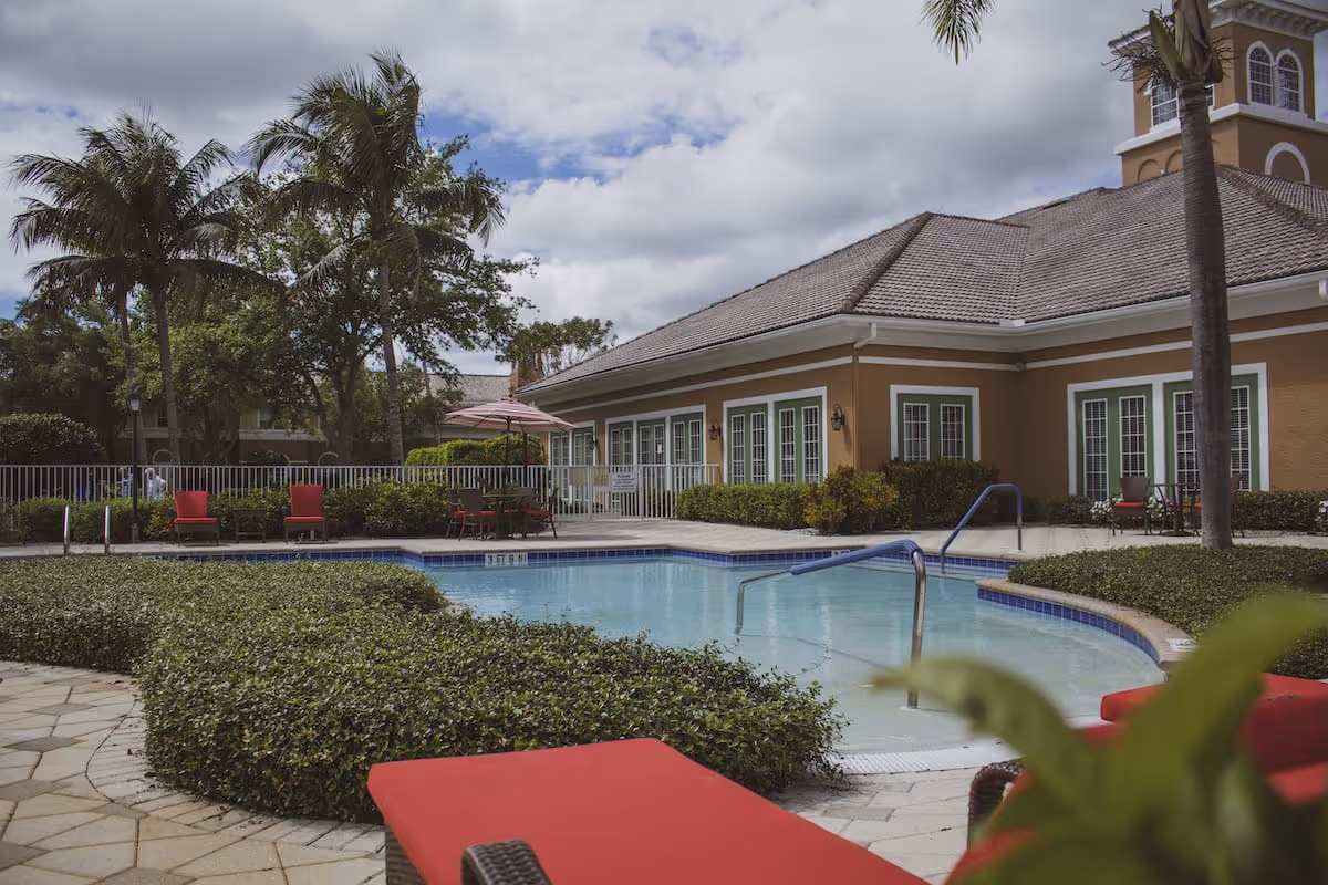Outdoor swimming pool area at Aston Gardens At Pelican Marsh with red lounge chairs, palm trees, and a building with multiple windows in the background under a partly cloudy sky.
