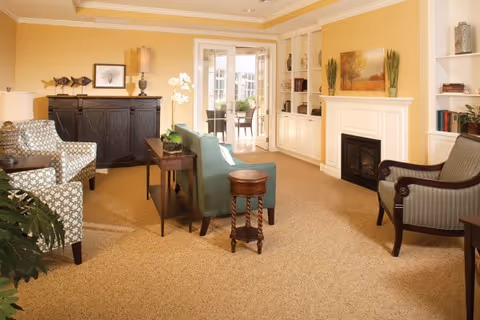 A cozy living room with beige carpet, a fireplace, and built-in white shelves. The room features a green upholstered sofa, patterned armchairs, a wooden side table, and decorative items including lamps, plants, and framed artwork. Sunlight streams in through glass doors leading to an outdoor area.