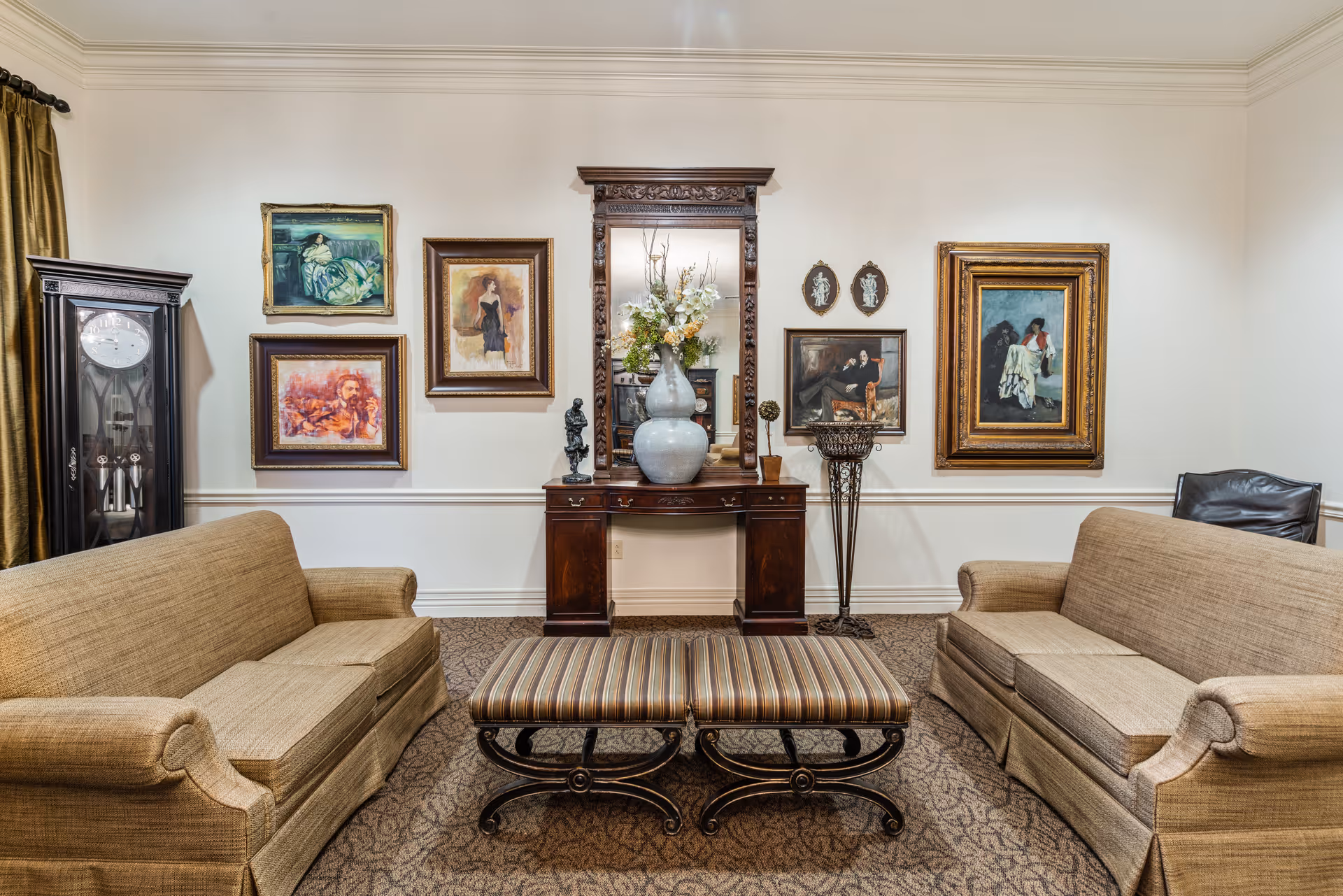 A cozy living room with two beige sofas facing each other, a pair of striped ottomans in the center, and a wooden console table with a large decorative mirror and flower arrangement. The walls are adorned with several framed paintings and artwork, and a tall grandfather clock stands in the corner.