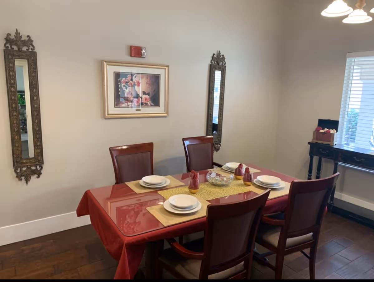 Dining room with a table set for four on a red tablecloth, wooden chairs, decorative mirrors and framed art on the wall.