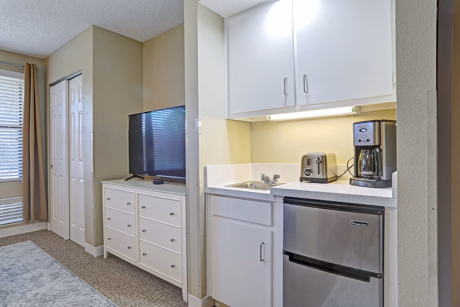 A small kitchenette area with white cabinets, a stainless steel mini refrigerator, a coffee maker, and a toaster. Adjacent to the kitchenette is a white dresser with a flat-screen TV on top. There is a window with blinds and beige curtains, and a closed closet with white sliding doors.