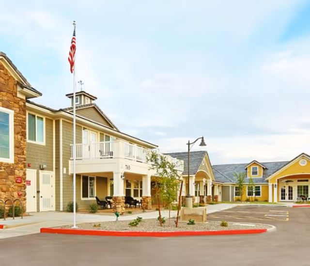 Exterior view of a senior living facility building with beige and yellow siding, a stone accent wall, and a balcony. There is an American flag on a flagpole in front, a parking area, small landscaped plants, and a street lamp.