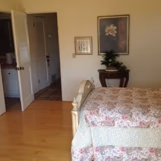 A bedroom with a bed covered in a floral quilt, a small wooden table with a plant and framed pictures on the wall, and an open door leading to a hallway and bathroom.