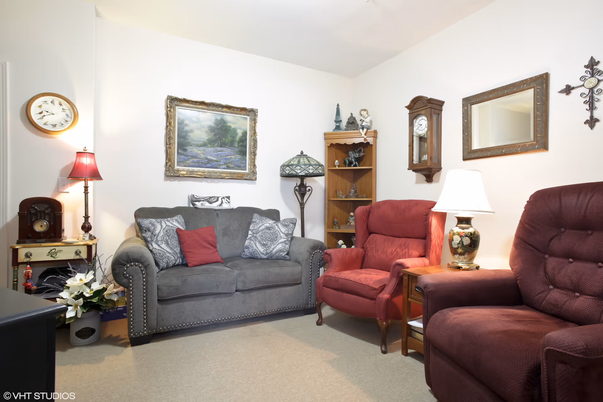 A cozy living room with a gray loveseat adorned with patterned and solid red pillows, two red armchairs, a wooden side table with a floral lamp, a wooden bookshelf with decorative items, a wall clock, a framed mirror, a wall-mounted clock, a floor lamp, a table lamp with a red shade, a small table with a vintage radio, and a framed landscape painting on the wall.
