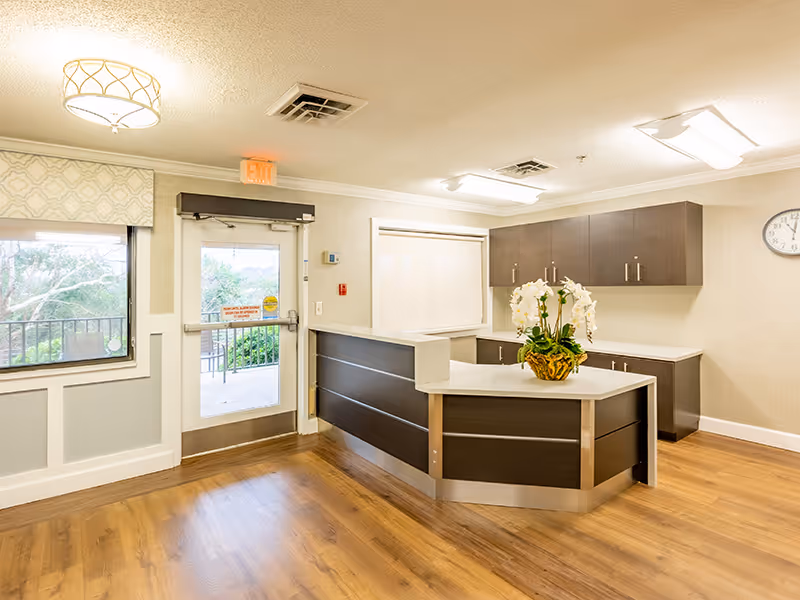 Reception area inside Brushy Creek Post Acute facility with a modern dark wood and white counter, a potted orchid plant on the counter, a window with a patterned valance, an exit door leading outside, and light wood flooring.