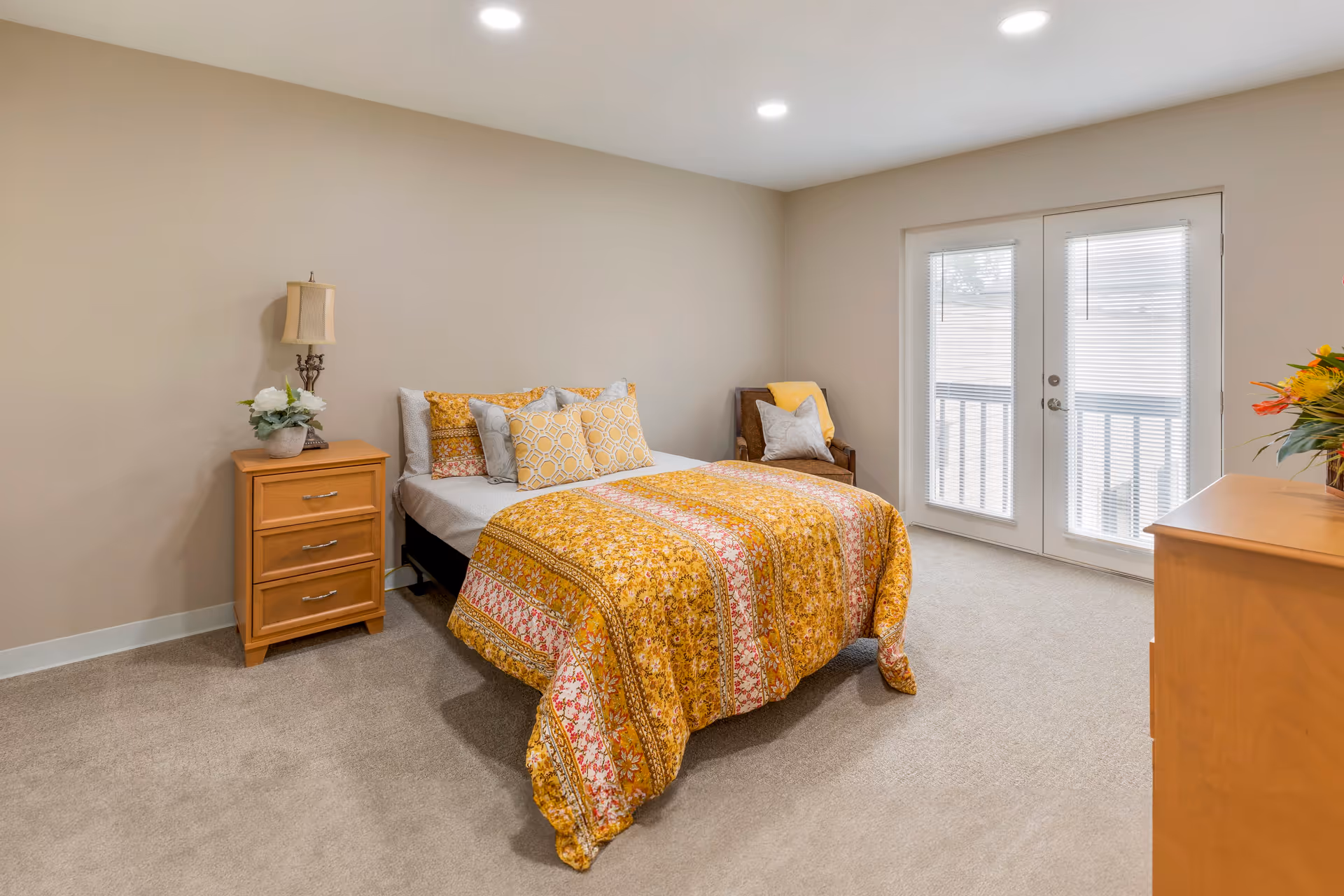 A cozy bedroom with a bed covered in a yellow and orange patterned quilt and multiple pillows. There is a wooden nightstand with a lamp and a small plant on the left side of the bed. A wooden dresser with a vase of flowers is on the right side of the room. The room has beige walls, carpeted floor, and double glass doors with blinds leading outside.