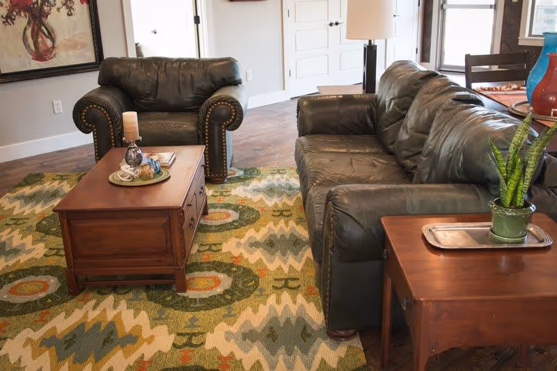 A cozy living room with a dark green leather sofa and matching armchair, a wooden coffee table with decorative items, a patterned area rug with green, yellow, and orange tones, a wooden side table with a potted plant, and a large window letting in natural light.