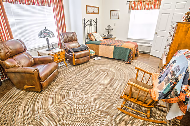 A cozy bedroom with a single bed covered in a patchwork quilt, two brown leather recliners, a wooden rocking chair with a colorful blanket draped over it, a braided oval rug on the floor, a wooden nightstand with a Tiffany-style lamp, and windows with striped curtains letting in natural light.