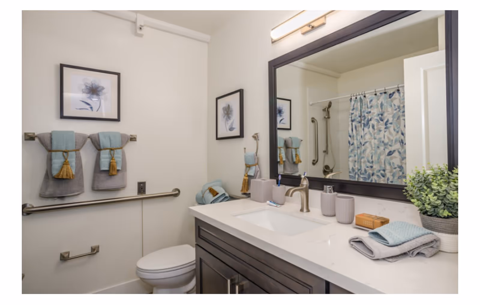 A clean and modern bathroom featuring a white toilet, a dark wood vanity with a white countertop, a large mirror with a dark frame, and a sink with a silver faucet. There are decorative towels in gray and light blue hanging on a towel rack and folded on the countertop. A small potted plant and soap are also on the countertop. The shower area has a curtain with a blue and white leaf pattern and a silver grab bar.