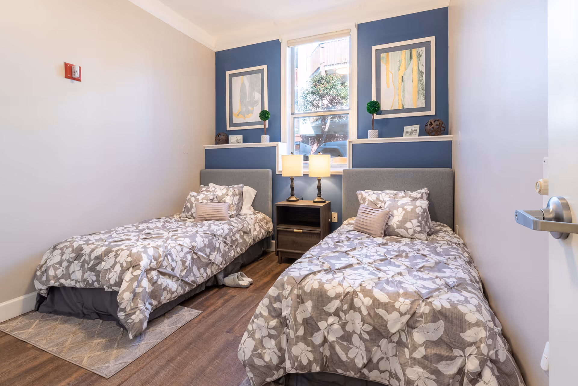 A bright bedroom with two twin beds featuring floral patterned bedding. Between the beds is a dark wooden nightstand with two lit table lamps. Above the beds are two framed abstract paintings and small decorative plants. A window in the center lets in natural light, showing a tree and building outside. The room has light-colored walls and wood flooring.