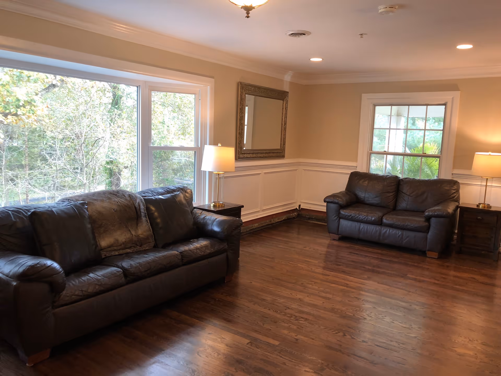 A living room with two dark brown leather sofas facing each other, a large window showing trees outside, two side tables each with a lamp, a large decorative mirror on the wall, and wooden flooring.
