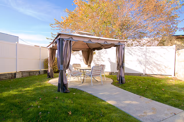 An outdoor gazebo with curtains over a round concrete pad holding a table and four chairs on a grassy yard next to a white fence.