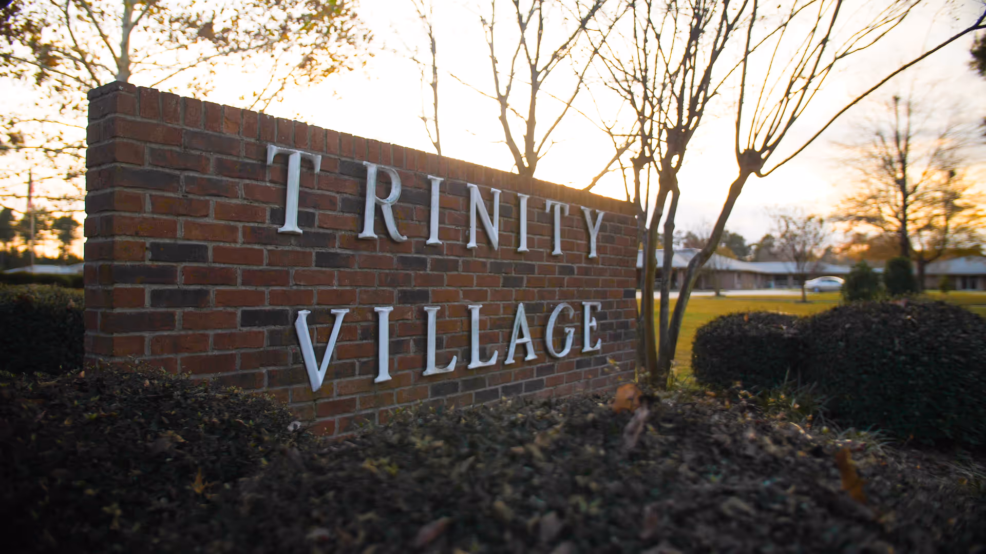 A brick sign with white letters spelling 'TRINITY VILLAGE' surrounded by bushes and trees, with a sunset sky in the background.