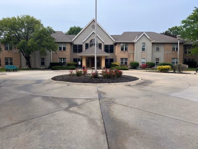 Front exterior view of The Northridge facility showing a two-story building with beige and light brown walls, multiple windows, a central entrance with a covered porch, a flagpole in front, and a circular driveway with a landscaped island containing flowers and mulch.