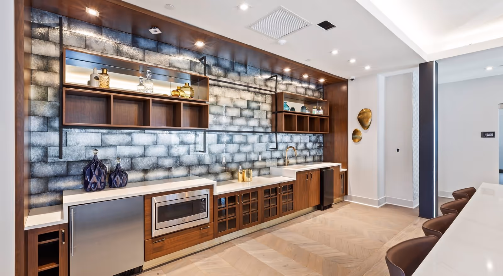 Modern kitchenette area with wooden cabinets, a microwave, a small refrigerator, a sink with a gold faucet, and open shelves displaying decorative glass bottles. The backsplash features a textured, metallic tile design. To the right, there is a white countertop with brown chairs lined up, and a wall with two gold decorative wall hangings. A sign indicating the fitness center is visible in the background.