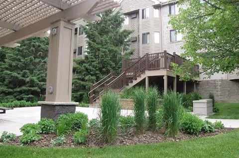 Outdoor area of a senior living facility with a covered patio, landscaped garden with green plants and grass, and a wooden staircase leading to an upper level of the building surrounded by trees.