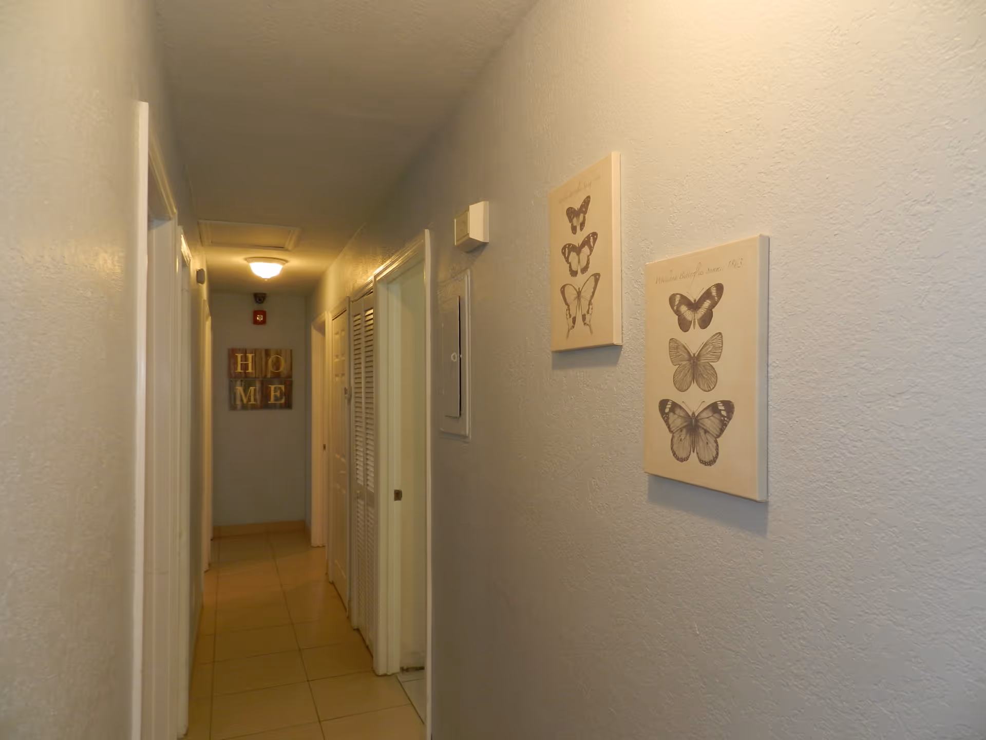 A narrow hallway with beige tiled floor and light-colored walls. On the right wall, there are two framed pictures of butterflies. At the end of the hallway, there is a wall decoration with the word 'HOME' in block letters. Several doors line the hallway on both sides, and a ceiling light fixture illuminates the space.