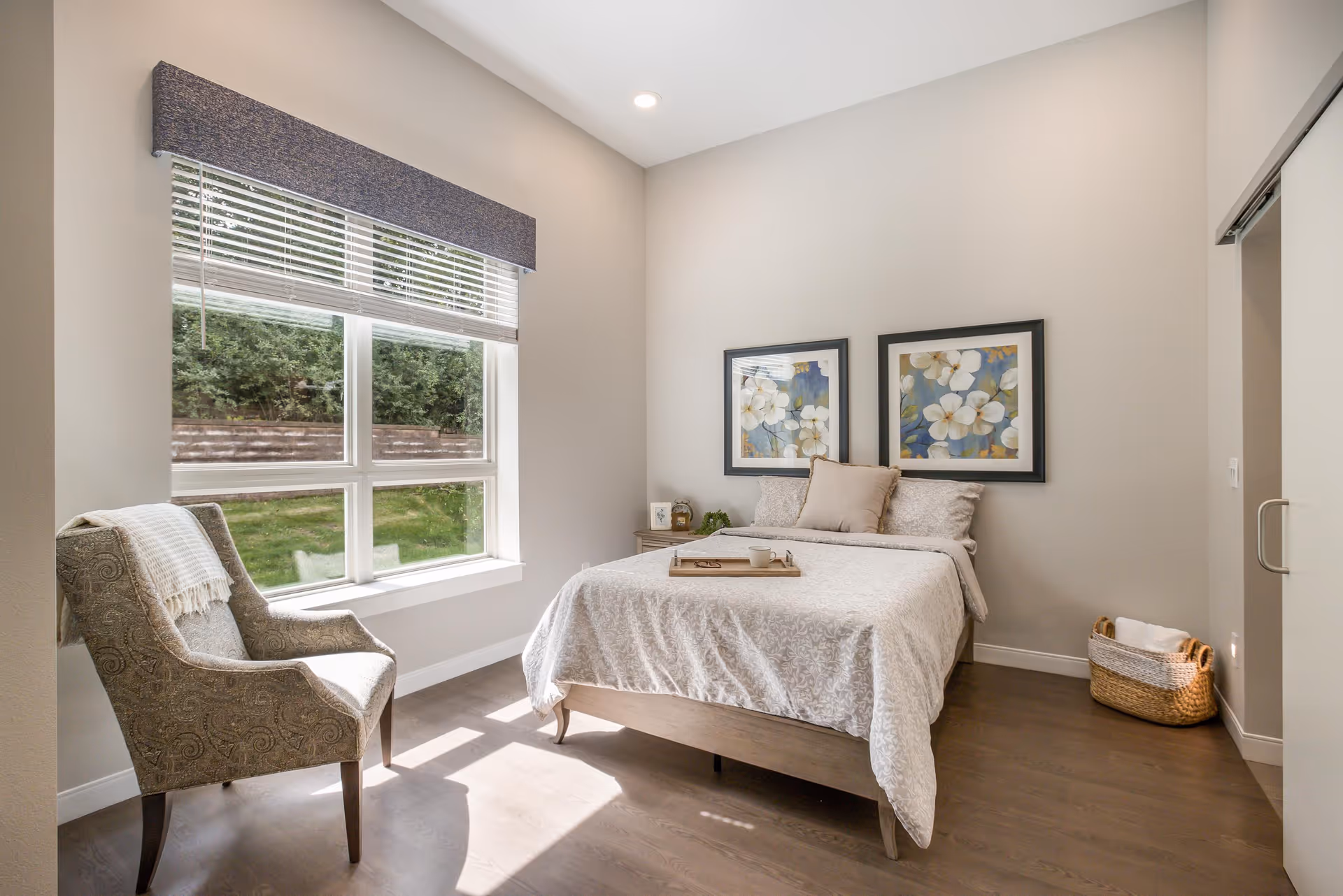 Sunlit bedroom with a bed topped by neutral bedding and floral artwork, an armchair by a large window, and a woven basket on the floor.