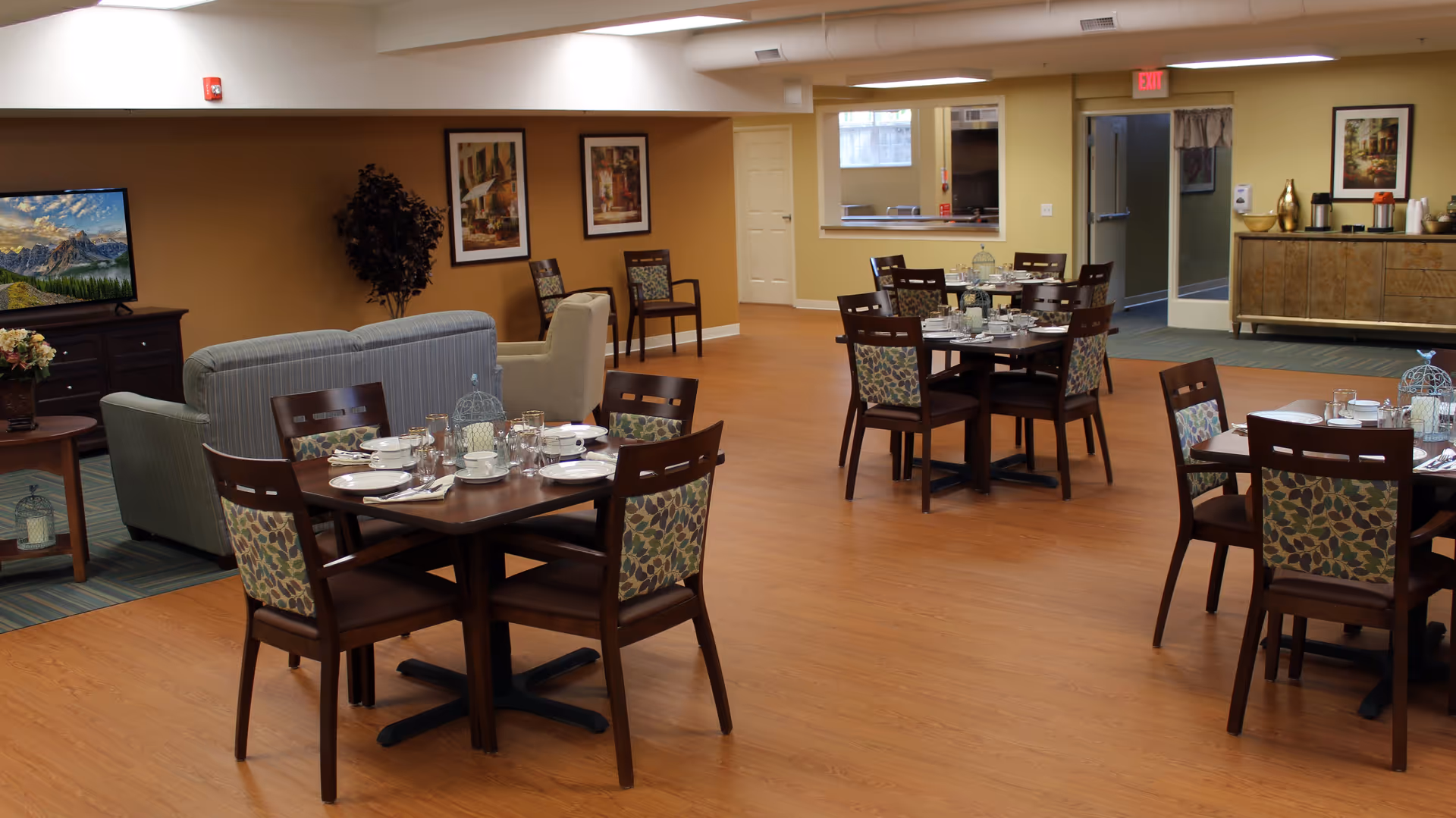 A dining area in a senior living facility with multiple wooden tables set with plates, cups, and silverware. The chairs have patterned upholstery. In the background, there is a seating area with couches and a TV displaying a mountain landscape. The walls are decorated with framed artwork, and there is a sideboard with coffee and other items near an open doorway leading to another room.