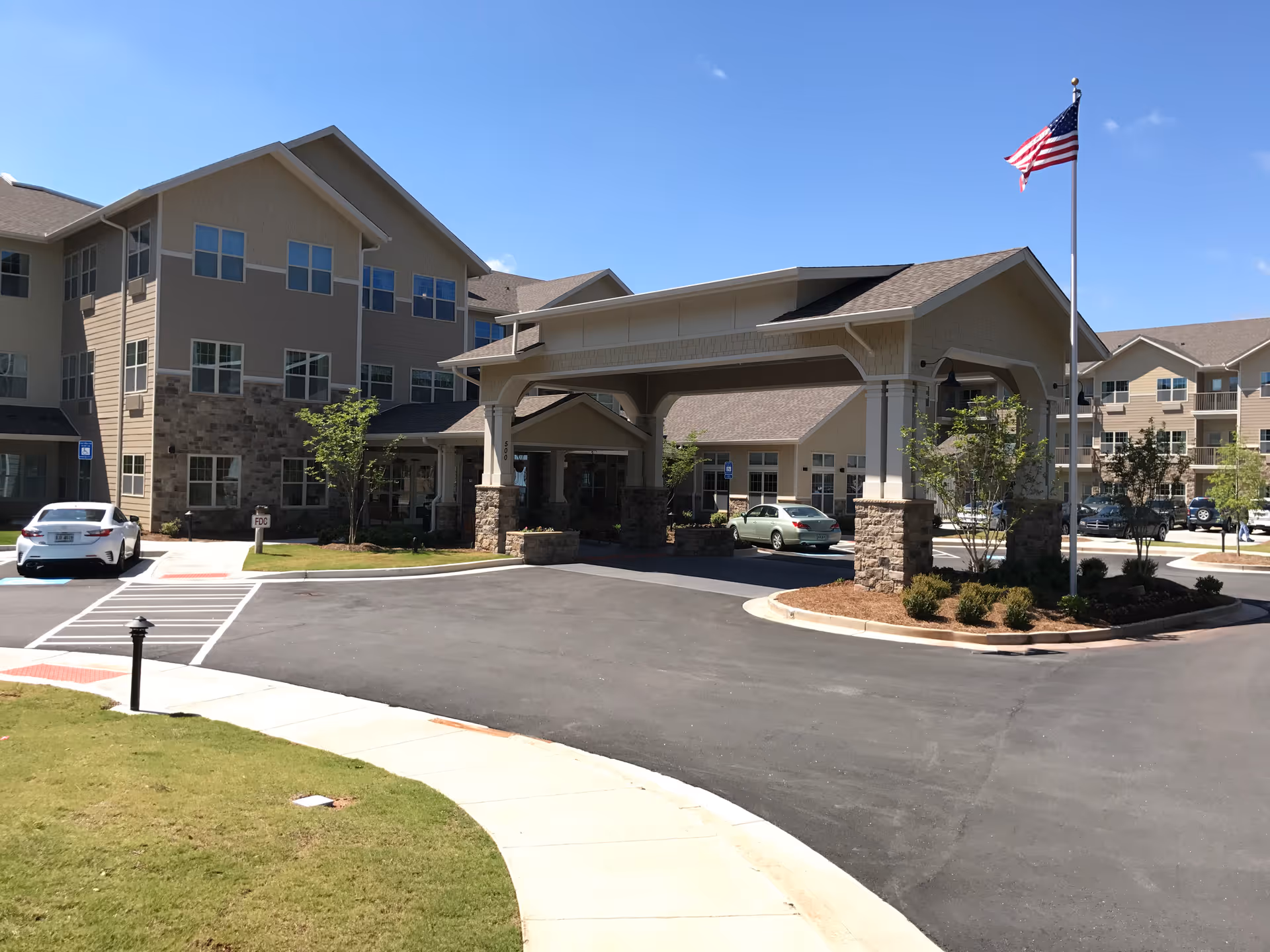 Exterior view of Sanford Estates Gracious Retirement Living facility showing a large covered entrance with stone pillars, an American flag on a flagpole, multiple parked cars, and a multi-story building under a clear blue sky.