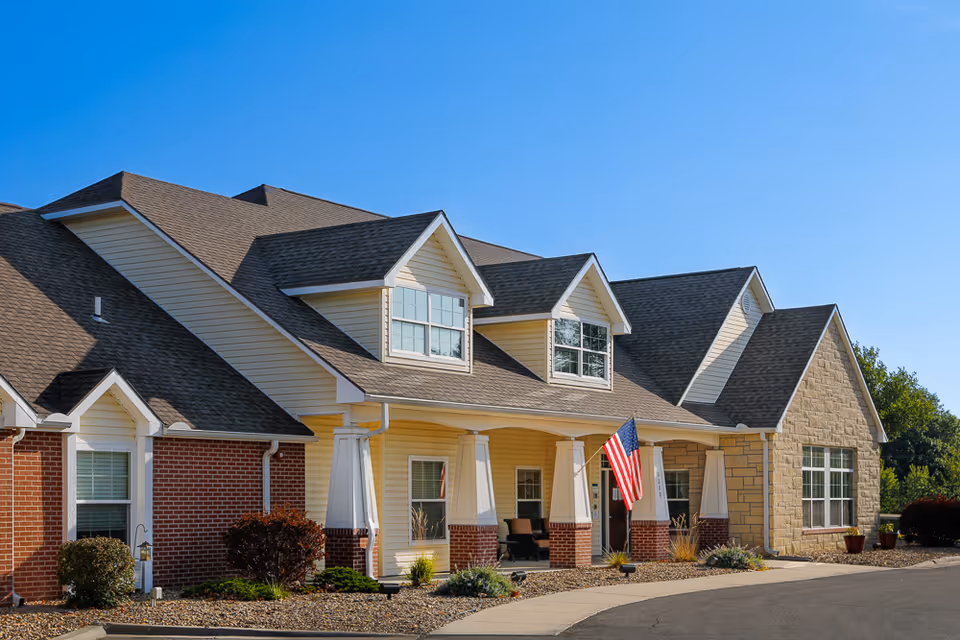Exterior view of a single-story assisted living facility building with a combination of brick, siding, and stone facade. The building features multiple gabled roofs, white columns supporting a covered porch, and an American flag displayed near the entrance. The sky is clear and blue.