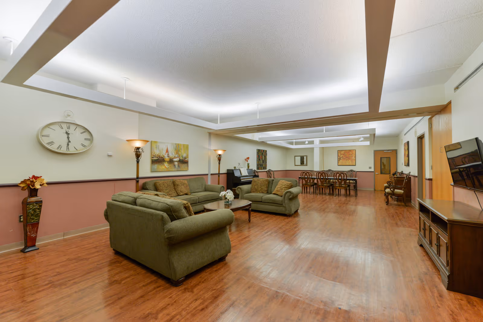 A spacious common area in a senior living facility with wooden flooring, two green sofas arranged around a coffee table, a piano against the wall, and a dining table with chairs in the background. The walls are decorated with paintings, a large clock, and floor lamps providing warm lighting.
