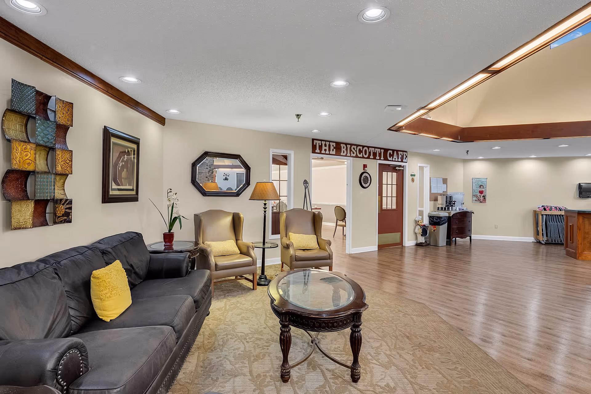 A cozy common area with a black leather sofa adorned with a yellow pillow, two beige armchairs with yellow cushions, a round glass-top wooden coffee table, and a floor lamp. The walls are decorated with a colorful metal wall art piece and a framed picture. In the background, there is a sign reading 'THE BISCOTTI CAFE' above a doorway leading to a dining area. The floor is wooden with a large beige rug under the seating area, and the ceiling has recessed lighting.