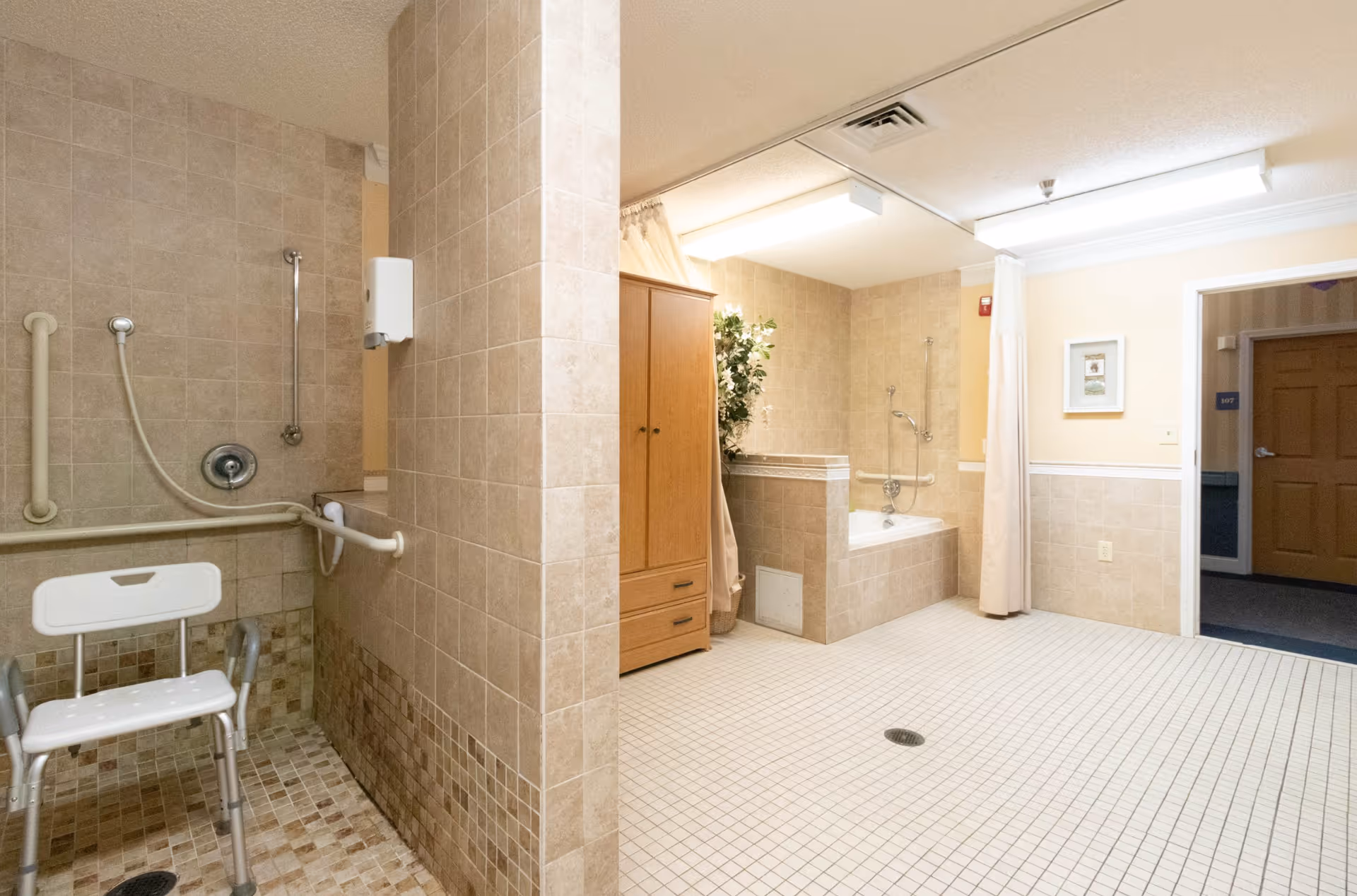 A spacious bathroom with beige tiled walls and floors. On the left side, there is a shower area with a white shower chair and grab bars. On the right side, there is a bathtub with grab bars and a shower curtain. A wooden cabinet and a potted plant are placed near the bathtub. The room is well-lit with ceiling lights, and there is an open doorway leading to a hallway with a wooden door labeled 107.