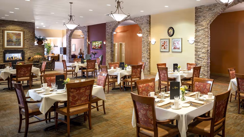 A spacious dining room with multiple tables covered in white tablecloths, each set with cups, glasses, plates, and menus. The room features stone archways, warm lighting, framed artwork on the walls, and a cozy fireplace in the background.