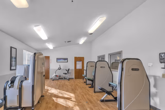 Bright exercise room with several seated fitness machines and a recumbent bike on a wood floor.