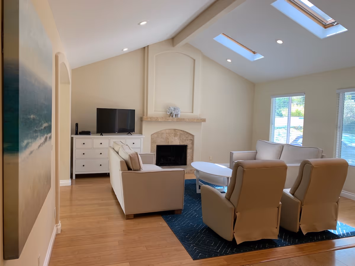 A bright living room with beige walls and wooden flooring featuring two beige armchairs and two beige sofas arranged around a white oval coffee table on a dark blue rug. There is a fireplace with a decorative elephant figurine on the mantel, a white dresser with a flat-screen TV on top, and two skylights in the ceiling letting in natural light. Two windows with blinds are visible on the right side.