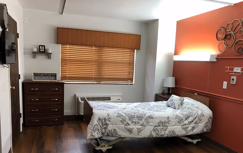 A single bed with a white and gray patterned bedspread in a room with wooden flooring. There is a wooden dresser with a small shelf above it holding a framed photo and a sign that reads 'Start each day with a grateful heart.' A window with closed wooden blinds is above an air conditioning unit. The wall behind the bed is painted a warm reddish color and decorated with circular metal wall art. A bedside table with a lamp is next to the bed.