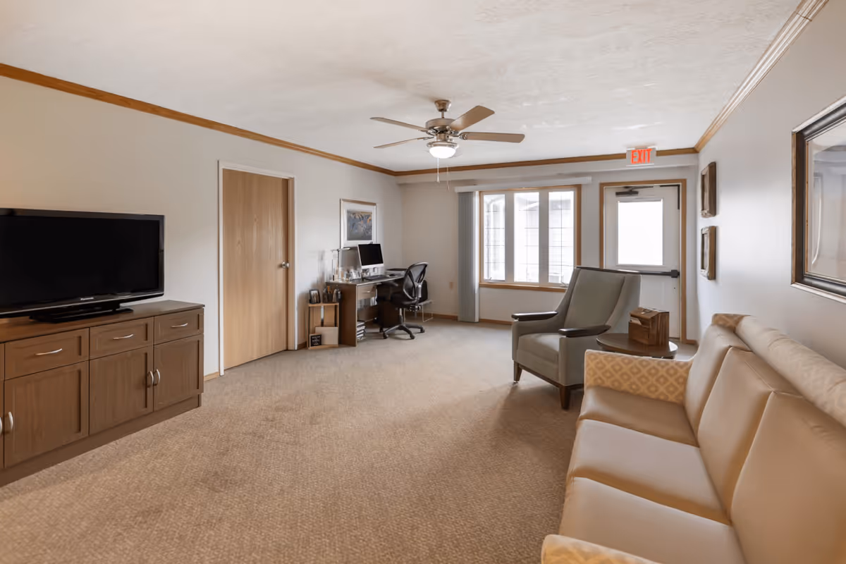 A spacious senior living room with beige carpet and light-colored walls trimmed with wood. The room features a tan leather sofa with patterned armrests, a gray armchair, a wooden side table, a wooden TV cabinet with a flat-screen TV, and a desk with a computer and office chair near a window. A ceiling fan with a light is mounted on the ceiling, and an exit door is visible at the far end of the room.