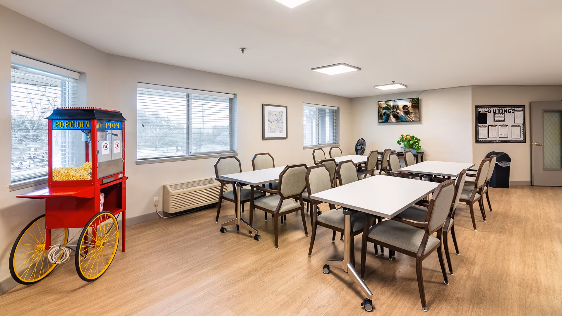 A bright dining room with several tables and chairs arranged neatly. There is a red popcorn machine on wheels filled with popcorn near the windows. The room has large windows with blinds, light-colored walls, wood flooring, a wall-mounted TV, a bulletin board, and a trash bin near the door.